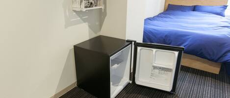 A hotel room with a bed, a small black refrigerator, and a pair of green slippers on the floor.