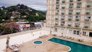 A hotel with a swimming pool, outdoor seating, and a view of a hillside with buildings.
