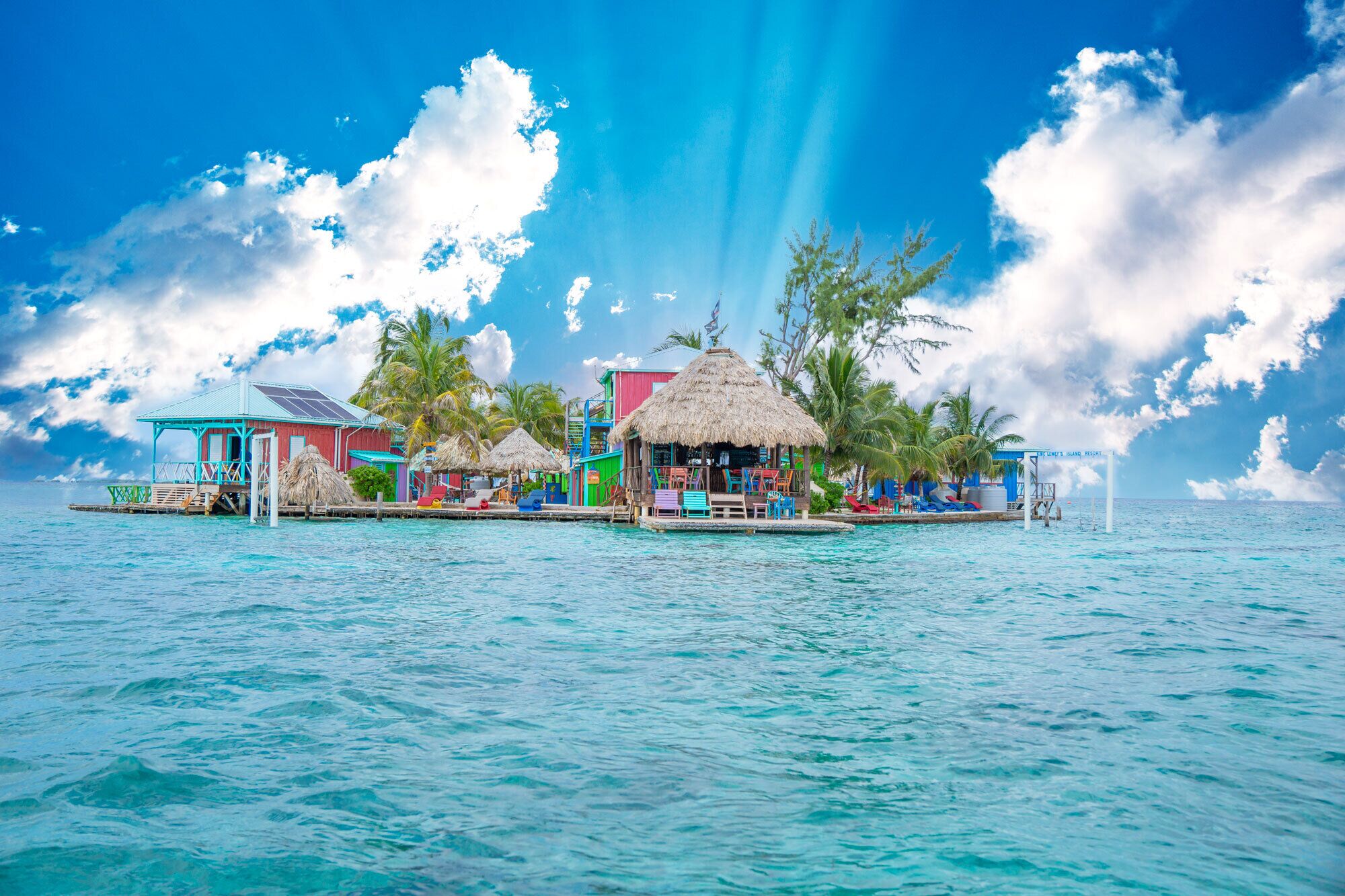A tropical resort with thatched-roof huts, colorful buildings, and a clear view of the ocean.