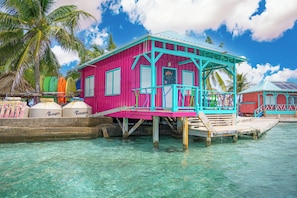 A pink and turquoise bungalow with a balcony, situated on stilts over clear water, surrounded by palm trees. - King Lewey's Island Resort (Placencia)
