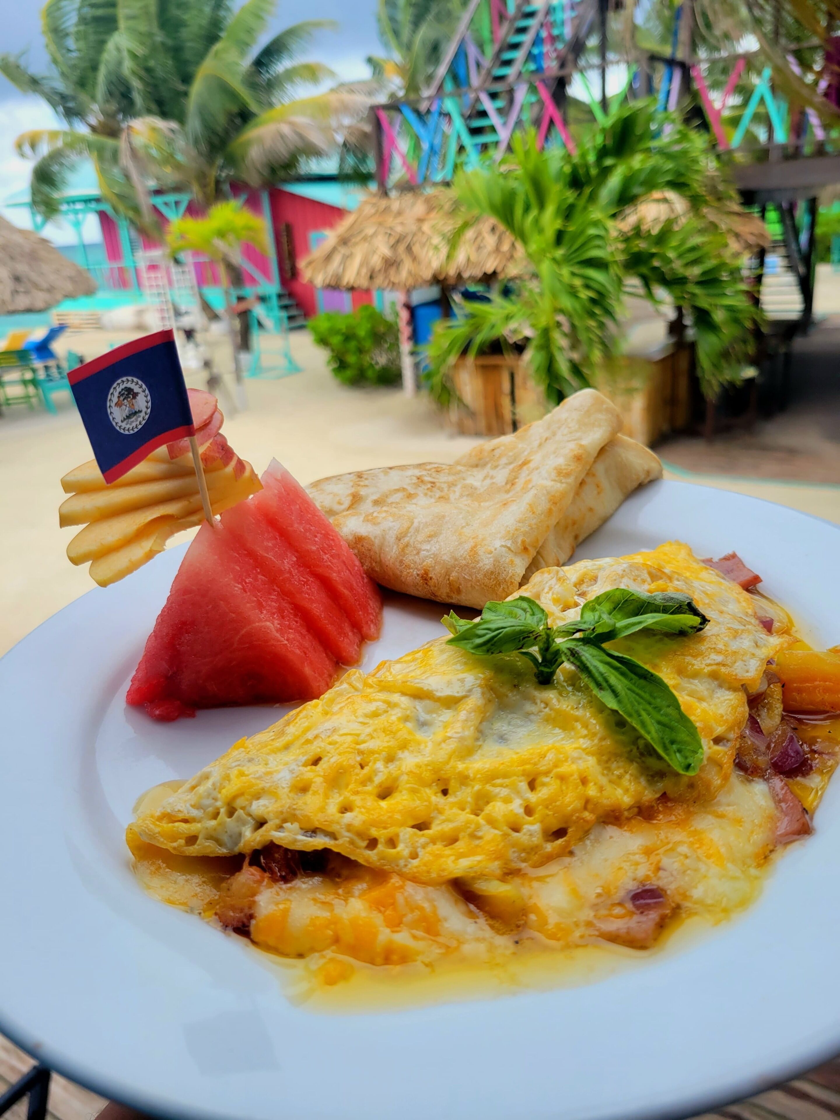 A plate with an omelette, ham, cheese, and a small flag, set against a beach backdrop with palm trees and colorful structures.