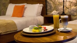 Uma cama bem arrumada com almofadas brancas e laranja, uma mesa de cabeceira com um abajur, e uma bandeja de café da manhã com frutas, iogurte e uma xícara de café.