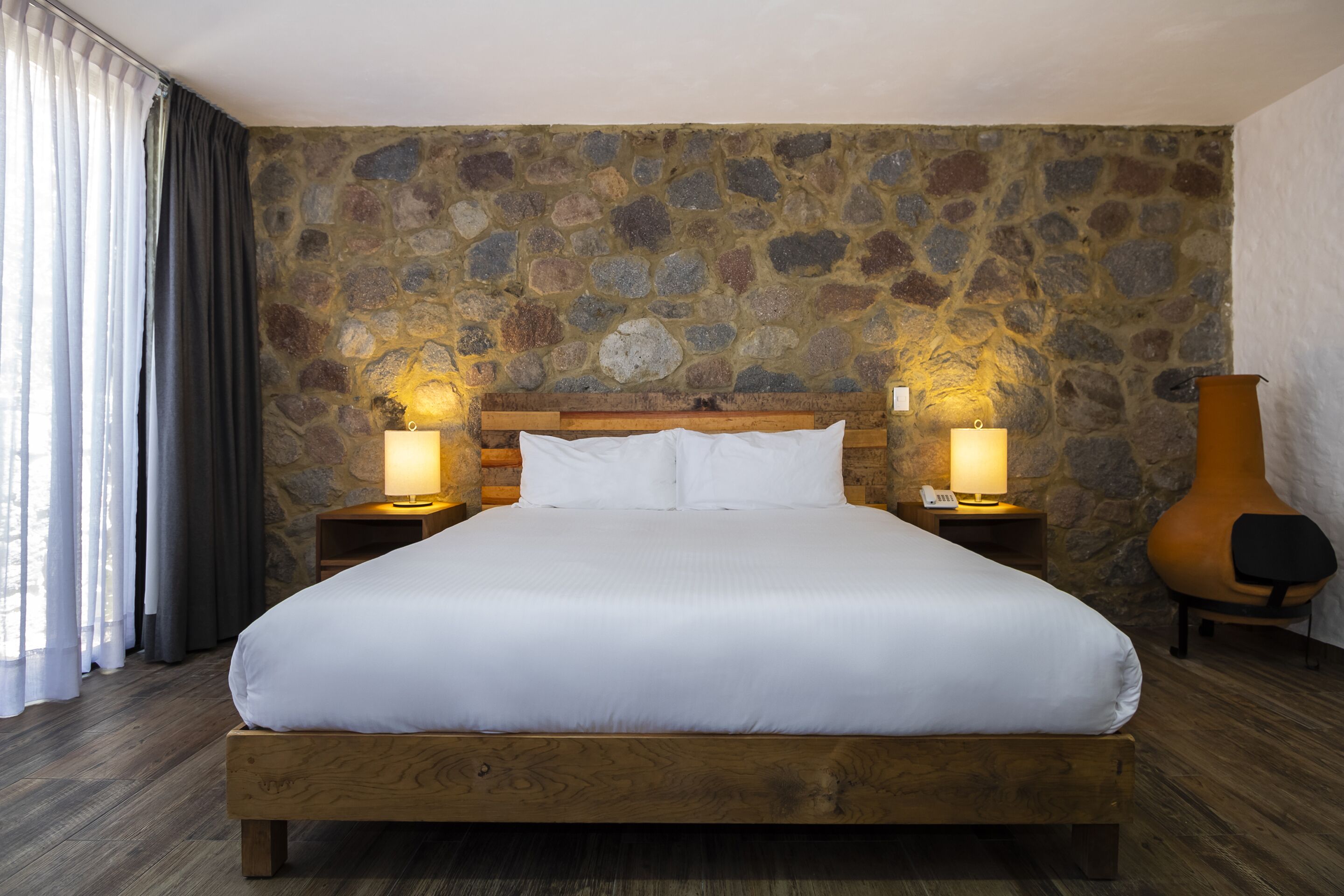 Una cama grande con ropa blanca, cabecera de madera y dos lámparas de noche. La habitación tiene una pared de piedra y piso de madera.