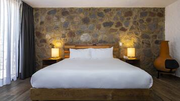 Una cama grande con ropa blanca, cabecera de madera y dos lámparas de noche. La habitación tiene una pared de piedra y piso de madera.