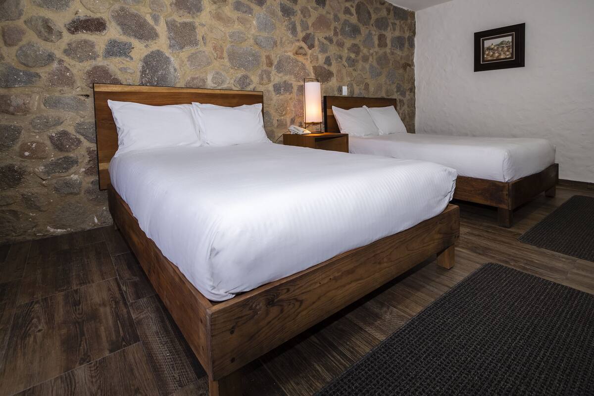 A double bed with a wooden frame and white bedding, a smaller single bed, a stone wall, and a framed picture on the wall.