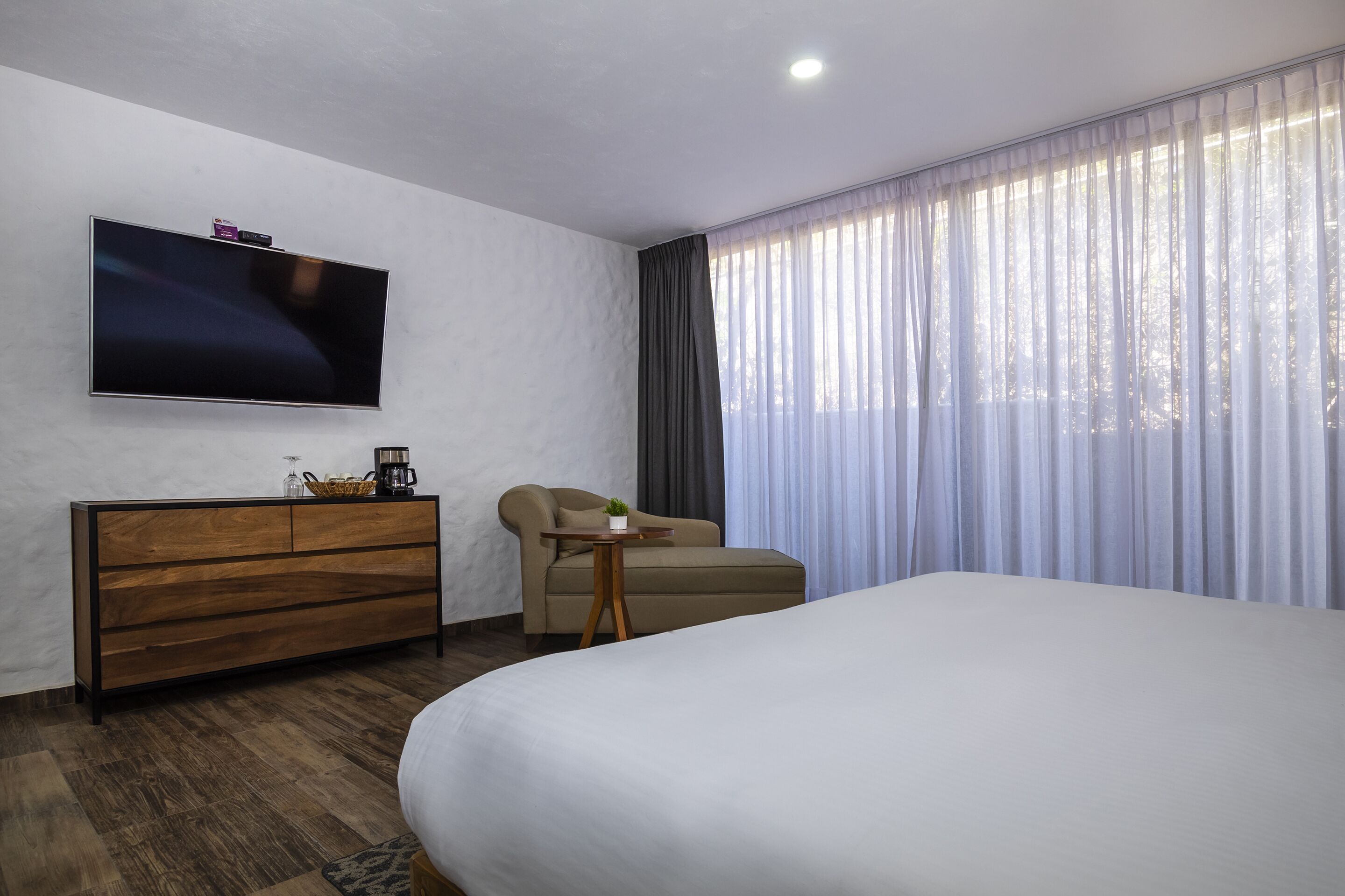 Ein Hotelzimmer mit einem großen Bett, einer Couch, einem an der Wand befestigten Fernseher, einer Holzkommode und einem Fenster mit durchsichtigen Vorhängen.
