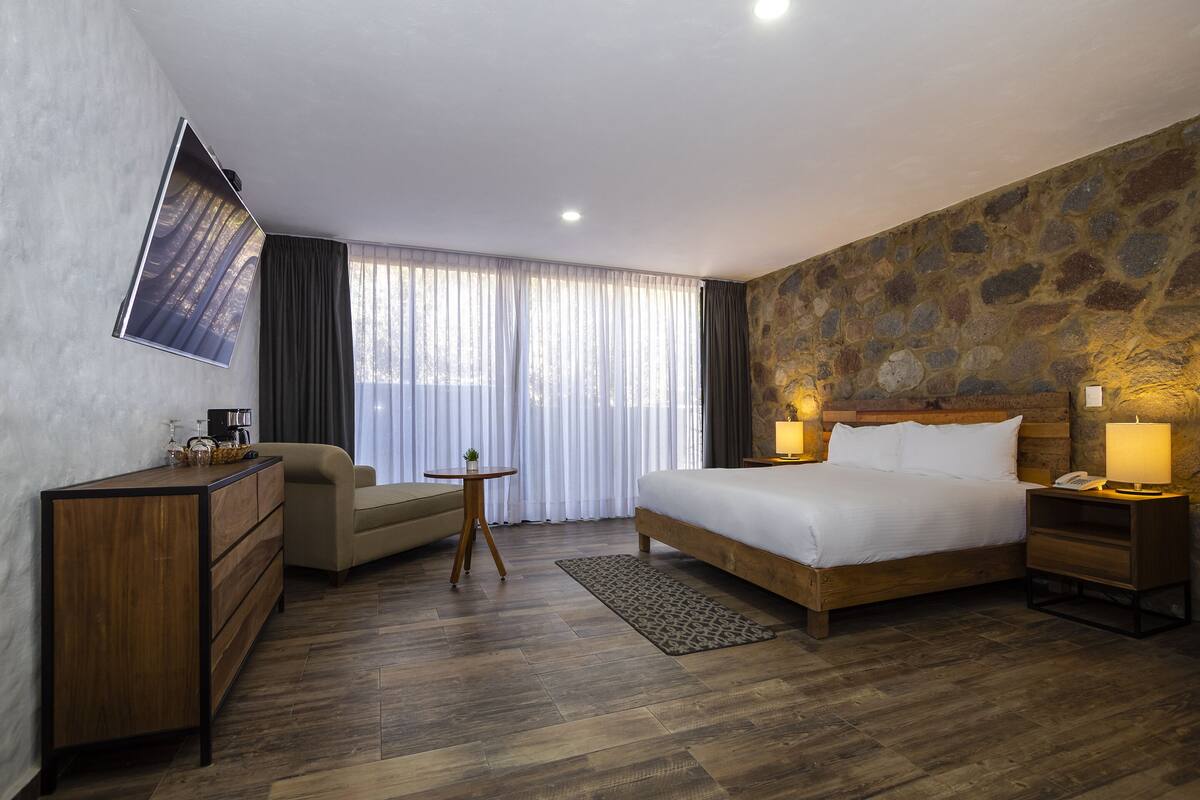 A hotel room with a wooden bed, a desk, a chair, a small table, and a TV mounted on the wall.