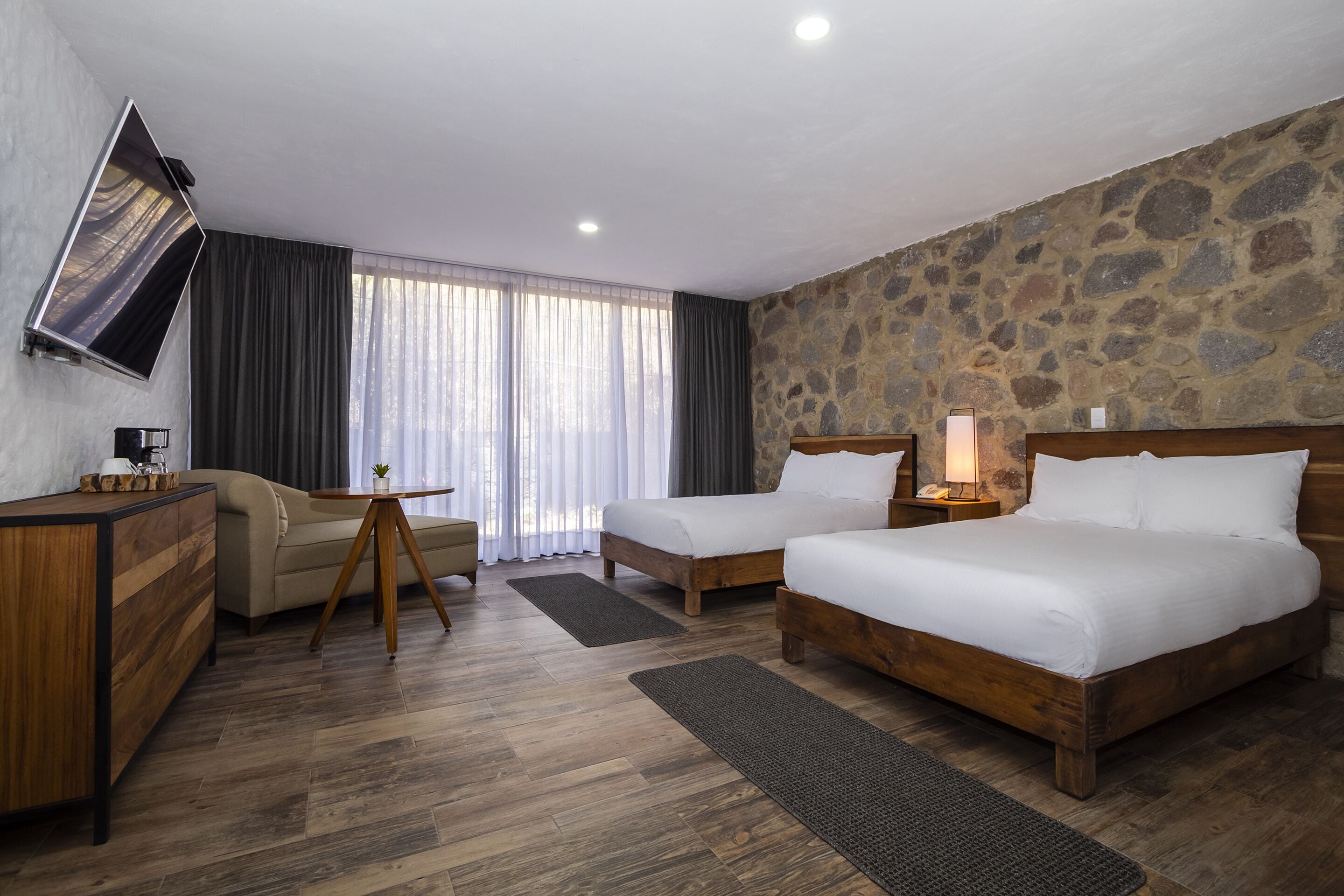 A hotel room with a stone wall, two beds, a desk, a TV, and a view of the outdoors through large windows.
