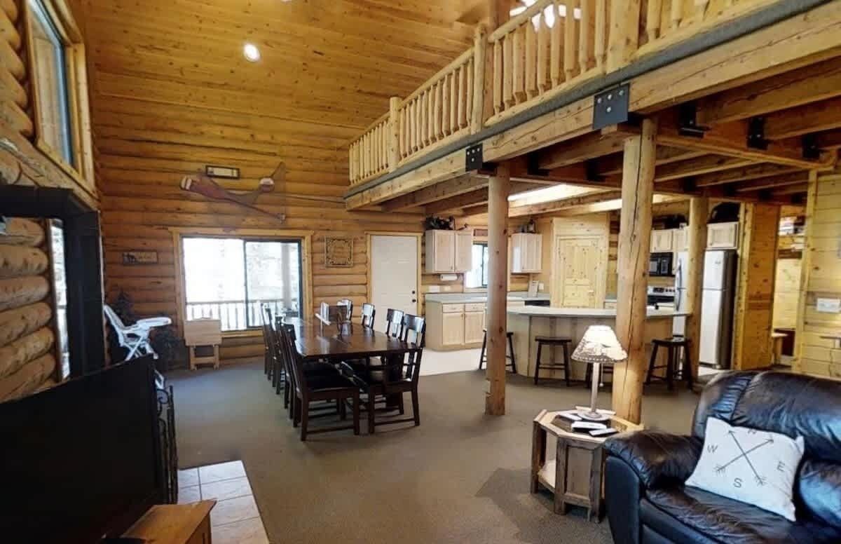 Interior de cabana de tronco com sala de jantar, cozinha e sala de estar com sofá e luminária.