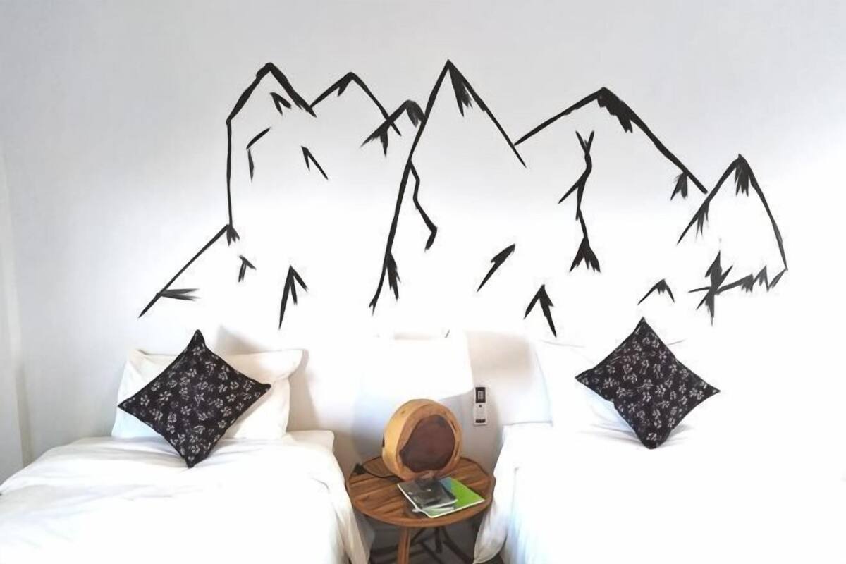 A hotel room with a bed, two pillows, a small round table with a book and a hat, and a wall with a mountain design.