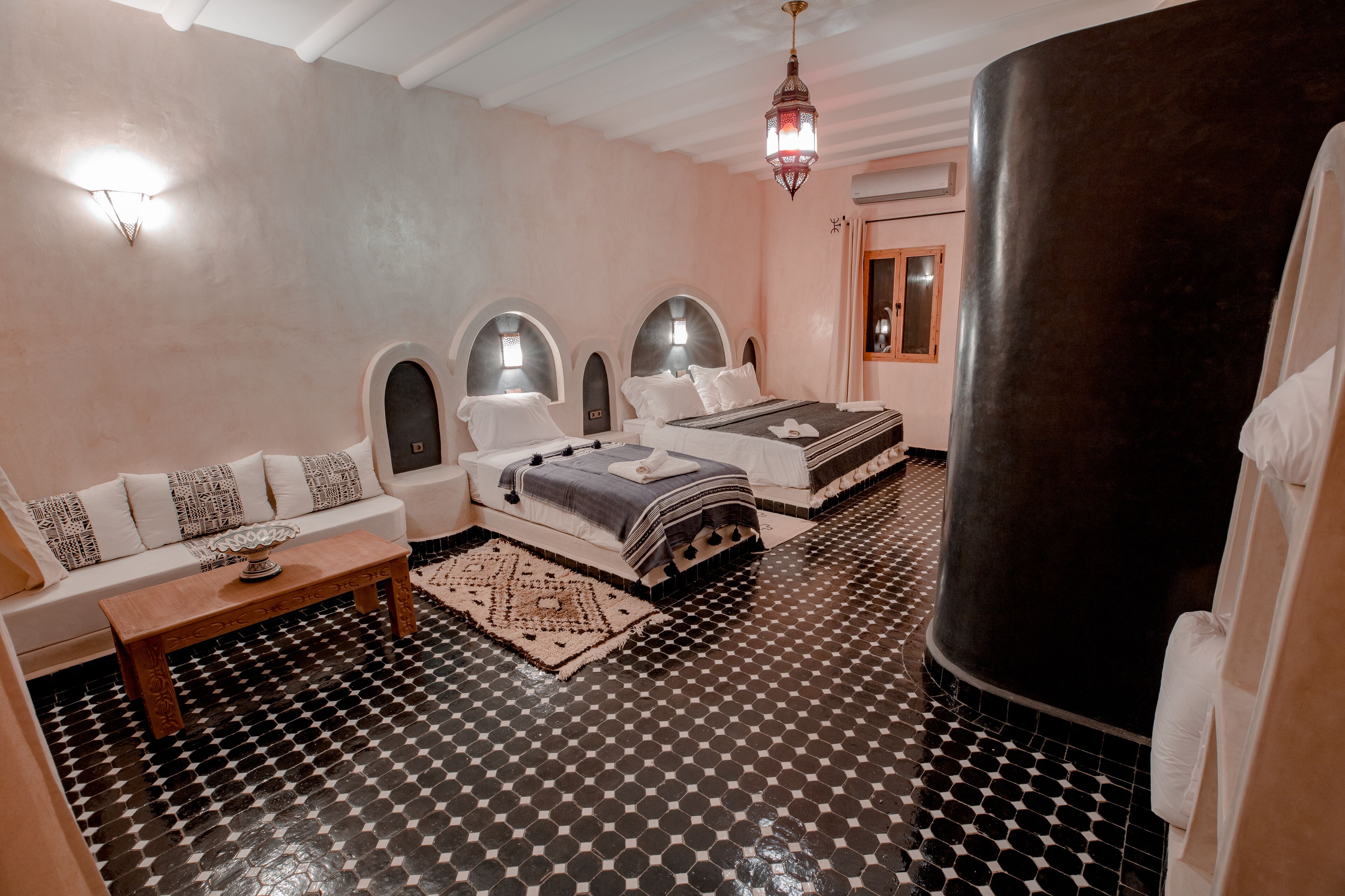 Habitació d'hotel amb terra de pissos, un sofà, dos llits, una taula de fusta i un objecte cilíndric gran.