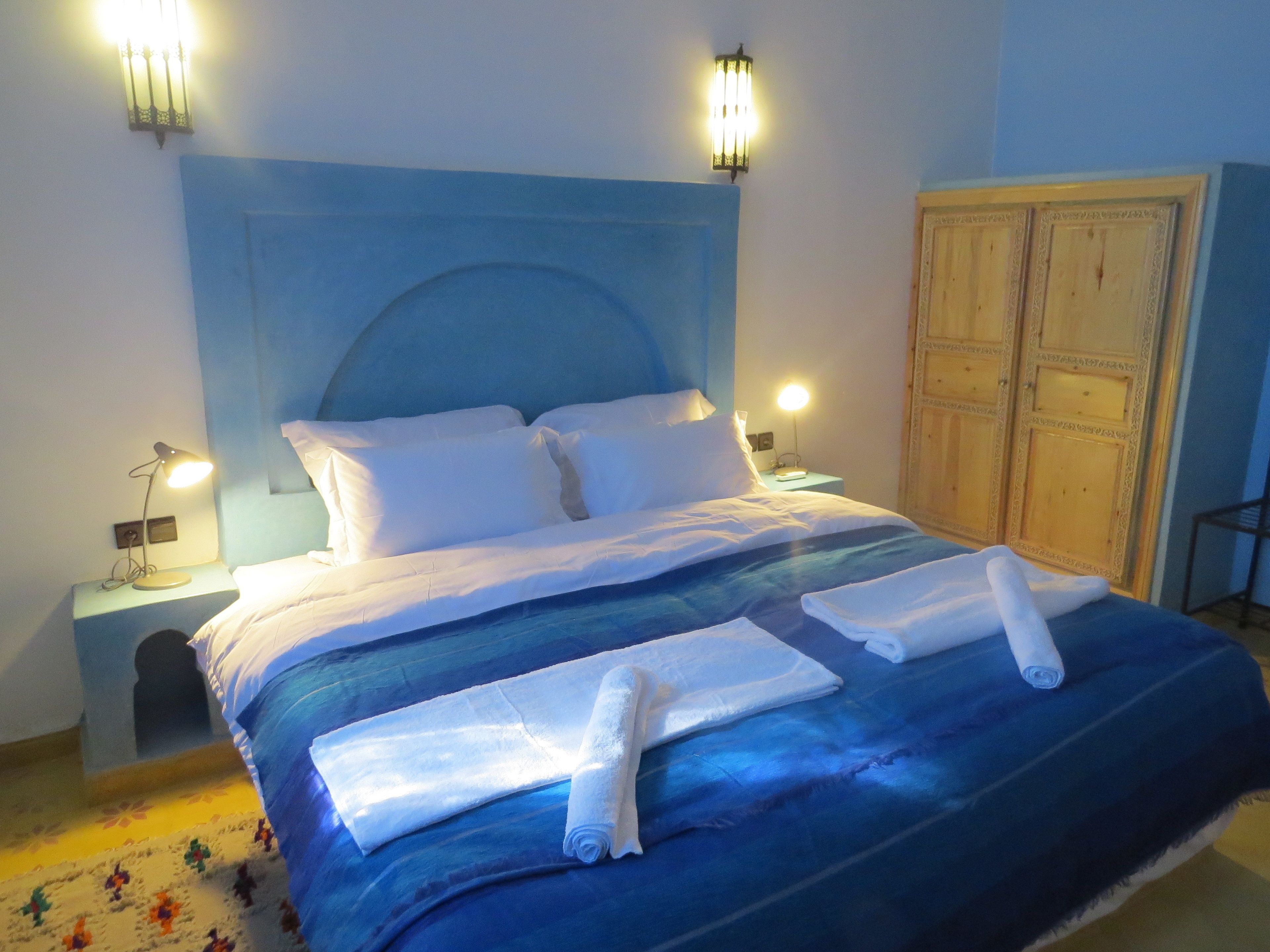 Una cama bien hecha con sábanas y toallas blancas, un cabecero azul y dos lámparas de noche.