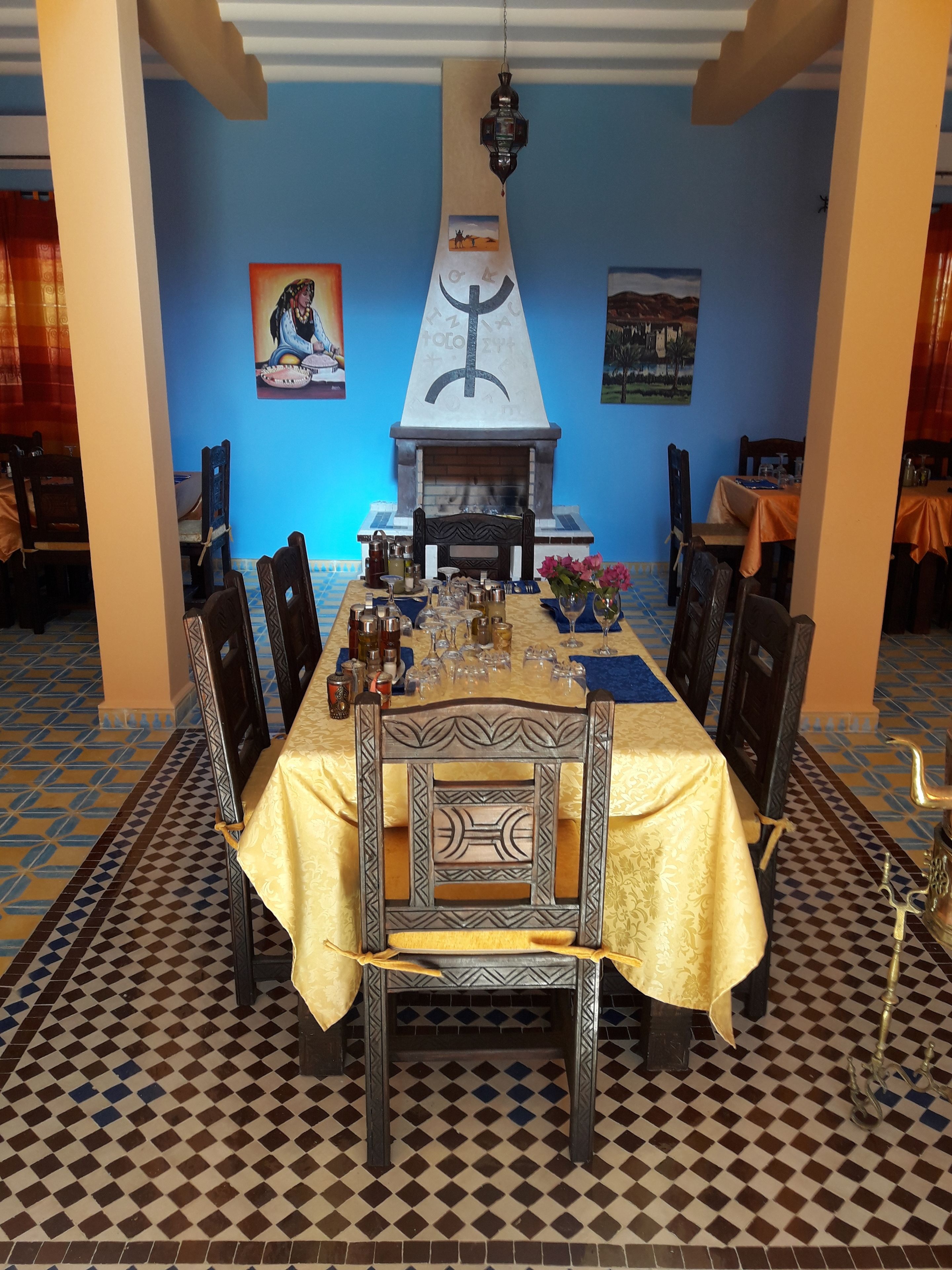 Une salle à manger avec une longue table dressée pour un repas, entourée de chaises. On aperçoit une cheminée au fond de la pièce, et un des murs est bleu et orné d’œuvres d’art.