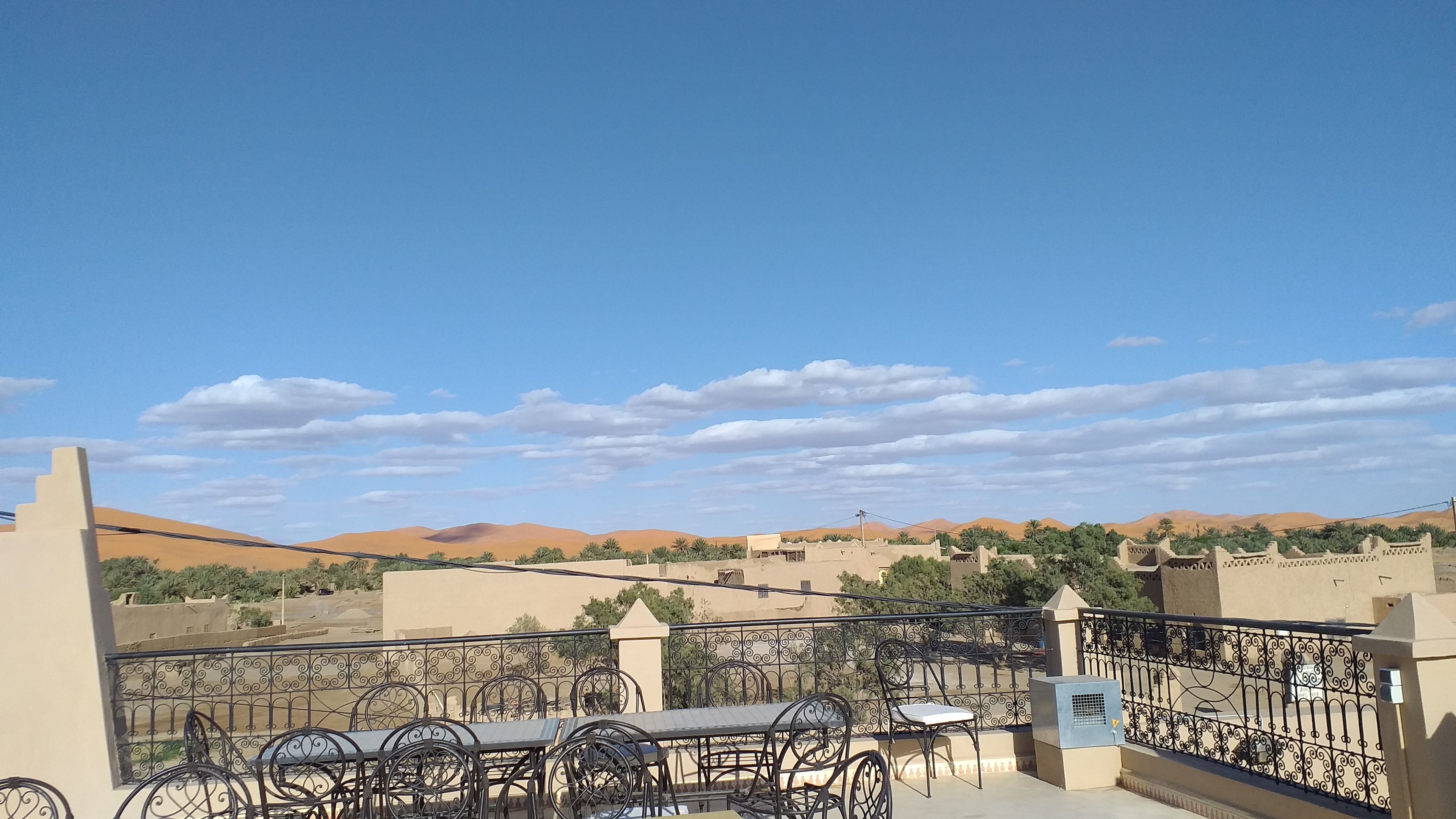 Varanda com mesa e cadeiras, com vista para uma paisagem desértica com dunas de areia e edifícios.