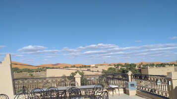 Varanda com mesa e cadeiras, com vista para uma paisagem desértica com dunas de areia e edifícios.