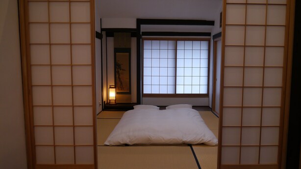 Une pièce de style japonais traditionnel, avec un sol recouvert de tatamis, un lit bas, des portes coulissantes en papier de riz (shoji) et une vue sur l’extérieur grâce à une grande fenêtre.