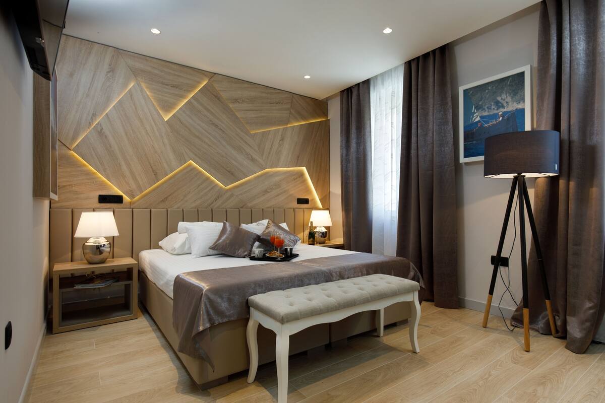 A modern hotel room with a large bed, bedside lamps, a bench, a desk lamp, and a framed picture on the wall.