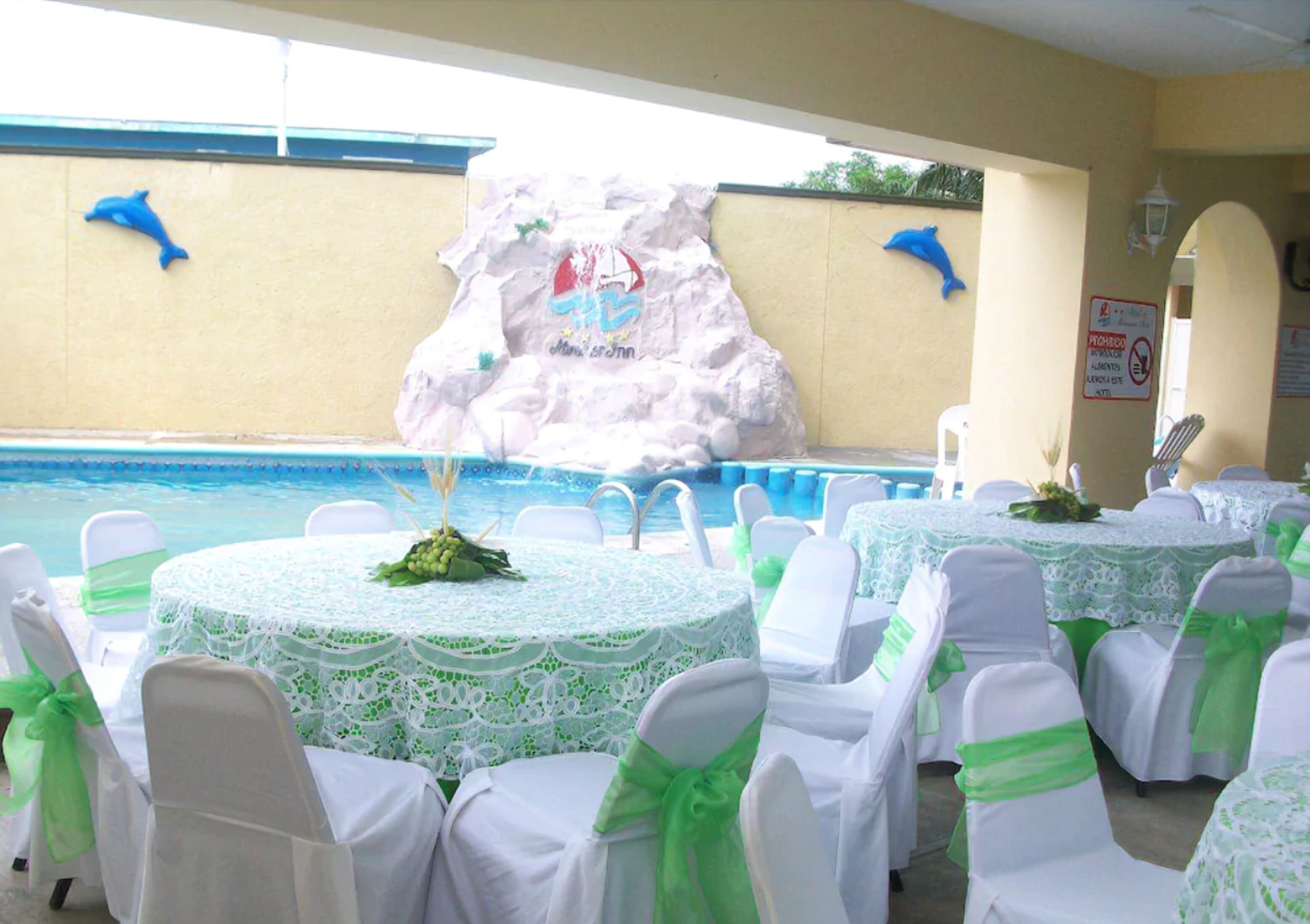 A poolside area with round tables covered in white lace tablecloths and chairs with green sashes. A decorative rock wall with a dolphin and a sign is visible in the background.