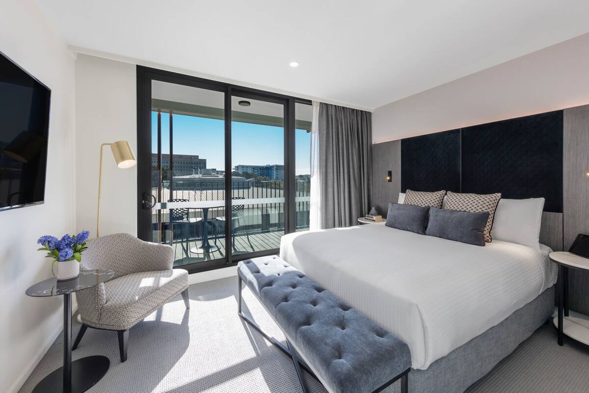 Quarto de hotel moderno com uma cama grande, área de estar e varanda com vista para a cidade.
