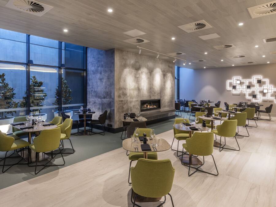 A modern restaurant with round tables and yellow chairs, a fireplace, and large windows.
