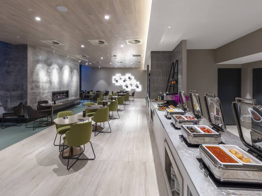 A modern dining area with multiple tables and chairs, a buffet with various food options, and a decorative light installation on the wall.