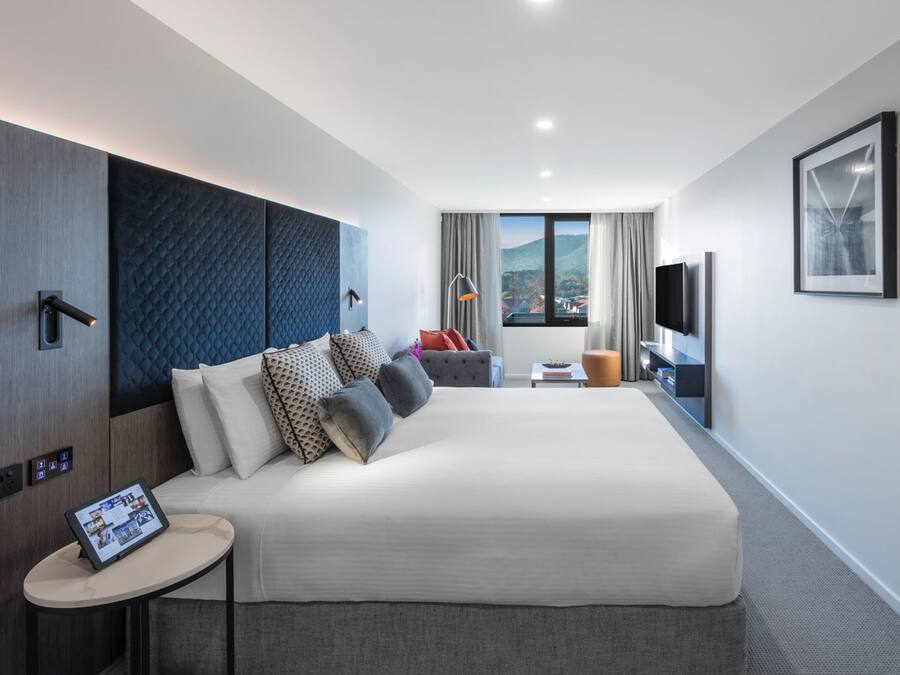 A modern hotel room with a large bed, a small side table, a TV, and a window with a city view.