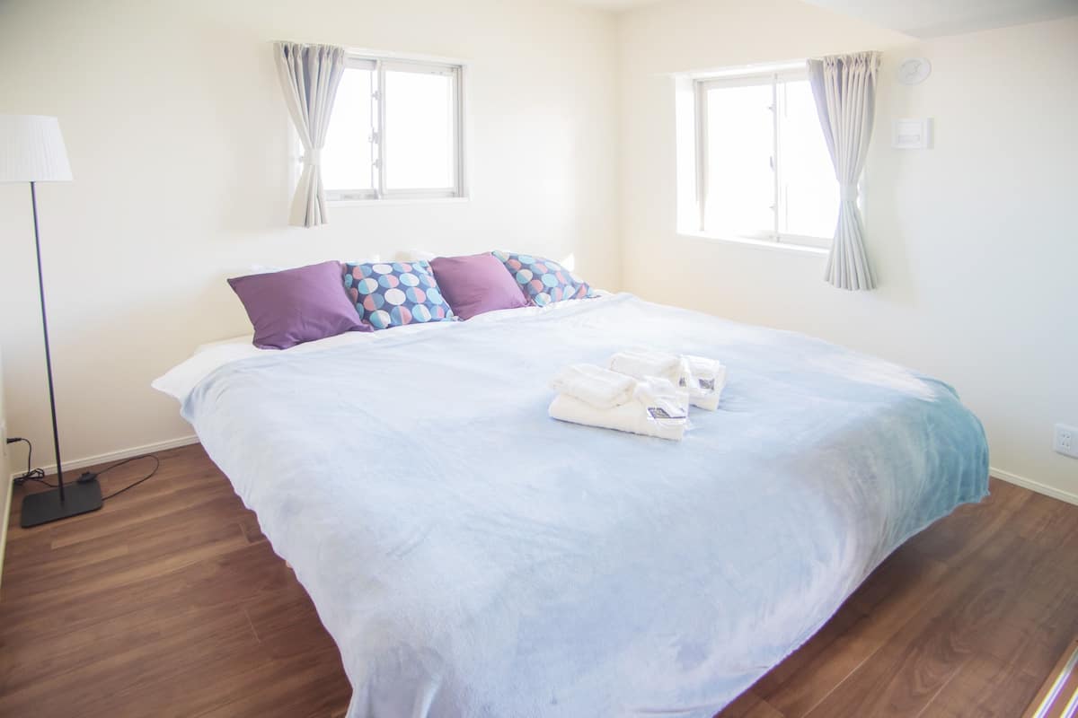 A neatly made bed with a blue and white duvet, two purple pillows, and a set of towels on the bed. There are two windows with curtains, a floor lamp, and a power outlet on the wall.