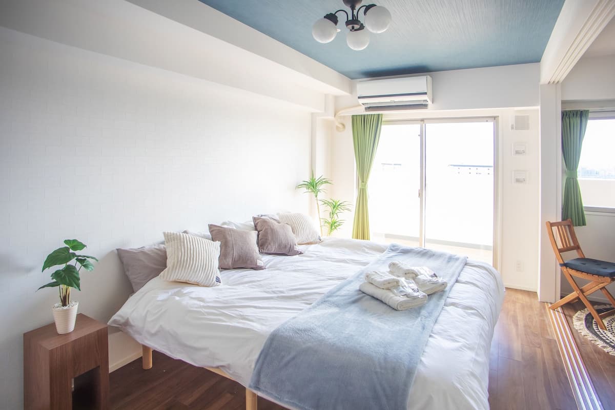 A bedroom with a bed, a bedside plant, a wooden nightstand, a window with curtains, and a view of the outside.
