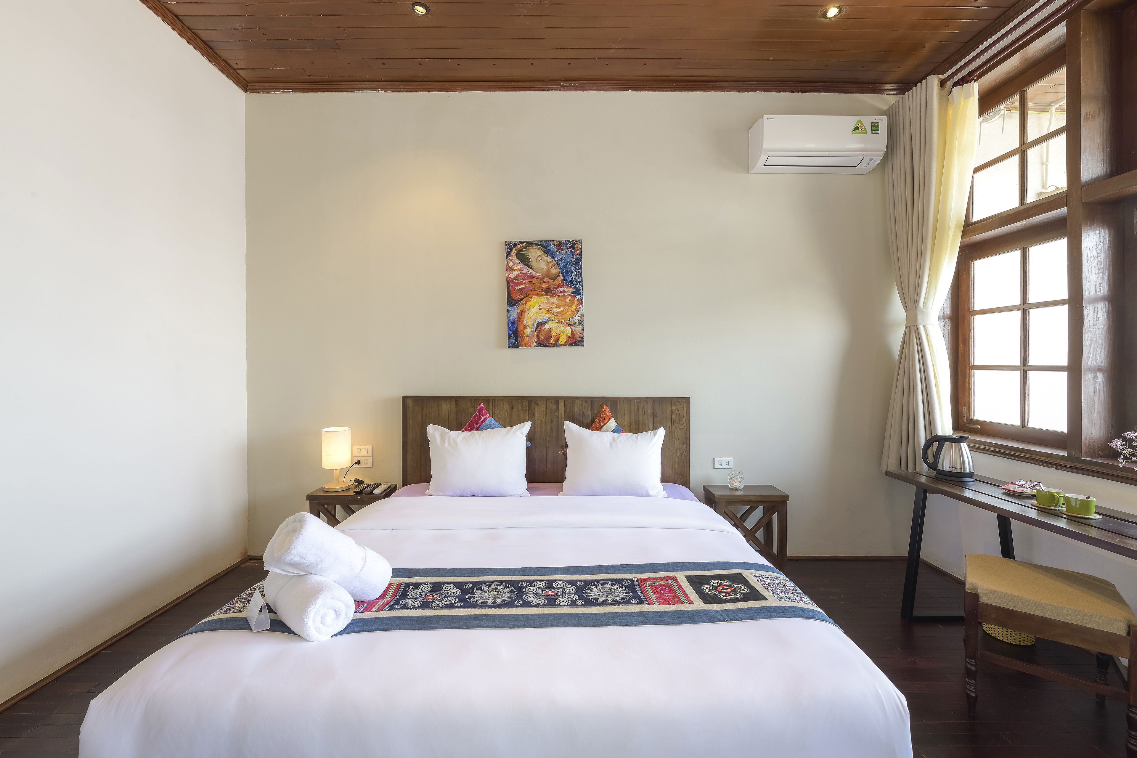 Una habitación de hotel con una cama grande, un cuadro en la pared, un escritorio con hervidor y una ventana con cortinas.