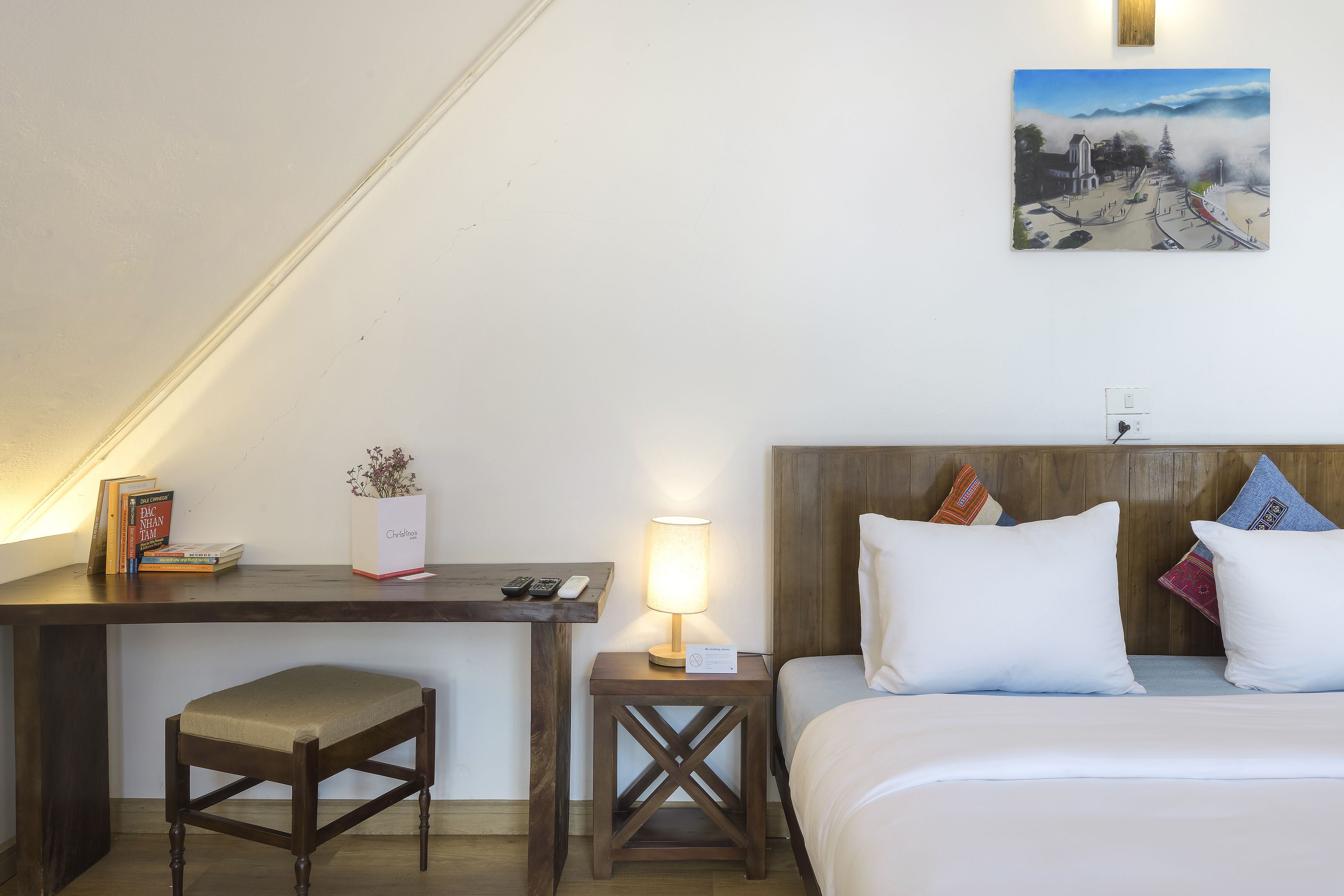 Habitación de hotel con cama, escritorio con silla, mesita de noche con lámpara, una pequeña mesa con libros y una planta, y un cuadro en la pared.