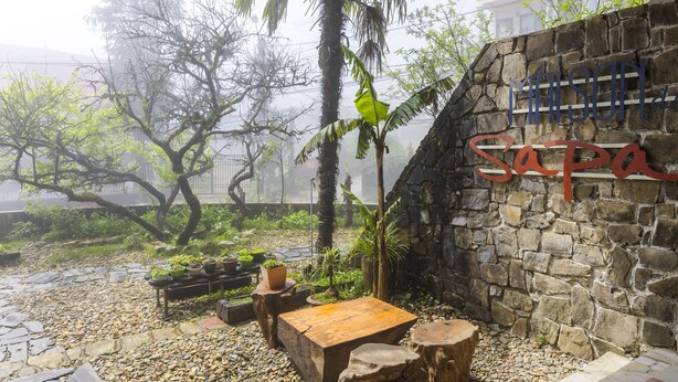 Un muro de piedra con el nombre "SAPA" y una zona de bancos de madera con plantas en macetas.