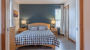 Ett sovrum med ett rutigt sängöverkast, två sänglampor och utsikt över naturen genom ett fönster.