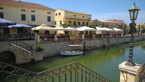 A riverside café with outdoor seating, stone walls, and a boat docked in the water. - Beautiful Apartments in a green and peaceful area near the beach on Corfu island (Lefkimmi)