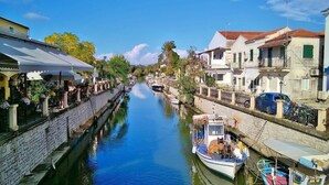A narrow canal flanked by buildings with balconies, a boat docked on the right, and a clear blue sky. - Beautiful Apartments in a green and peaceful area near the beach on Corfu island (Lefkimmi)