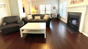 A living room with a dark wood floor, a white coffee table, a sofa with patterned cushions, a patterned armchair, a desk with a chair, a fireplace, and a ceiling fan. - University townhome,  available for long-term. (Charlotte)