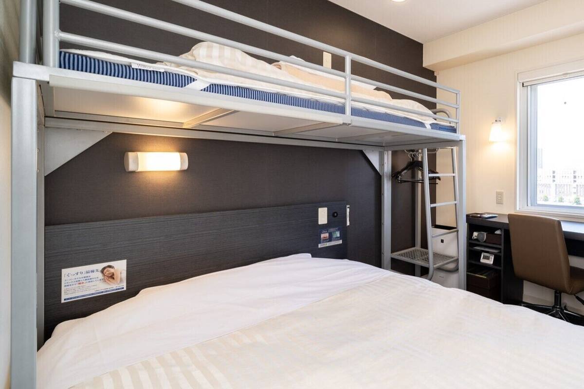 Een slaapkamer met een stapelbed, bureau en stoel, een raam en een wandplank.