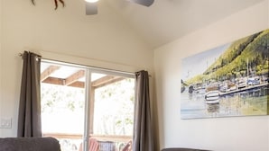 Ein Wohnzimmer mit einem grauen Sofa, einem Glastisch mit Krabben, einem Deckenventilator und einem Gemälde eines Hafens.