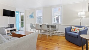 Ein Wohnzimmer mit grauem Sofa, blauem Sessel, Holztisch, Fernseher, Esstisch mit Stühlen und einem Fenster mit Jalousien.