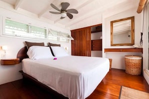 A bedroom with a bed, wooden floor, ceiling fan, window blinds, wardrobe, mirror, and a wicker basket.