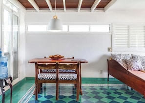 A room with a wooden table and chairs, a bed with patterned pillows, a water dispenser, and a window with blinds.