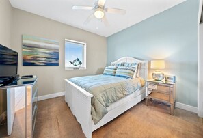 A bedroom with a bed, a desk, a ceiling fan, a window with blinds, and artwork on the wall.