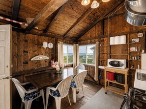 A rustic wooden interior with a dining table, chairs, a kitchen area with a microwave, and a view of the outdoors.