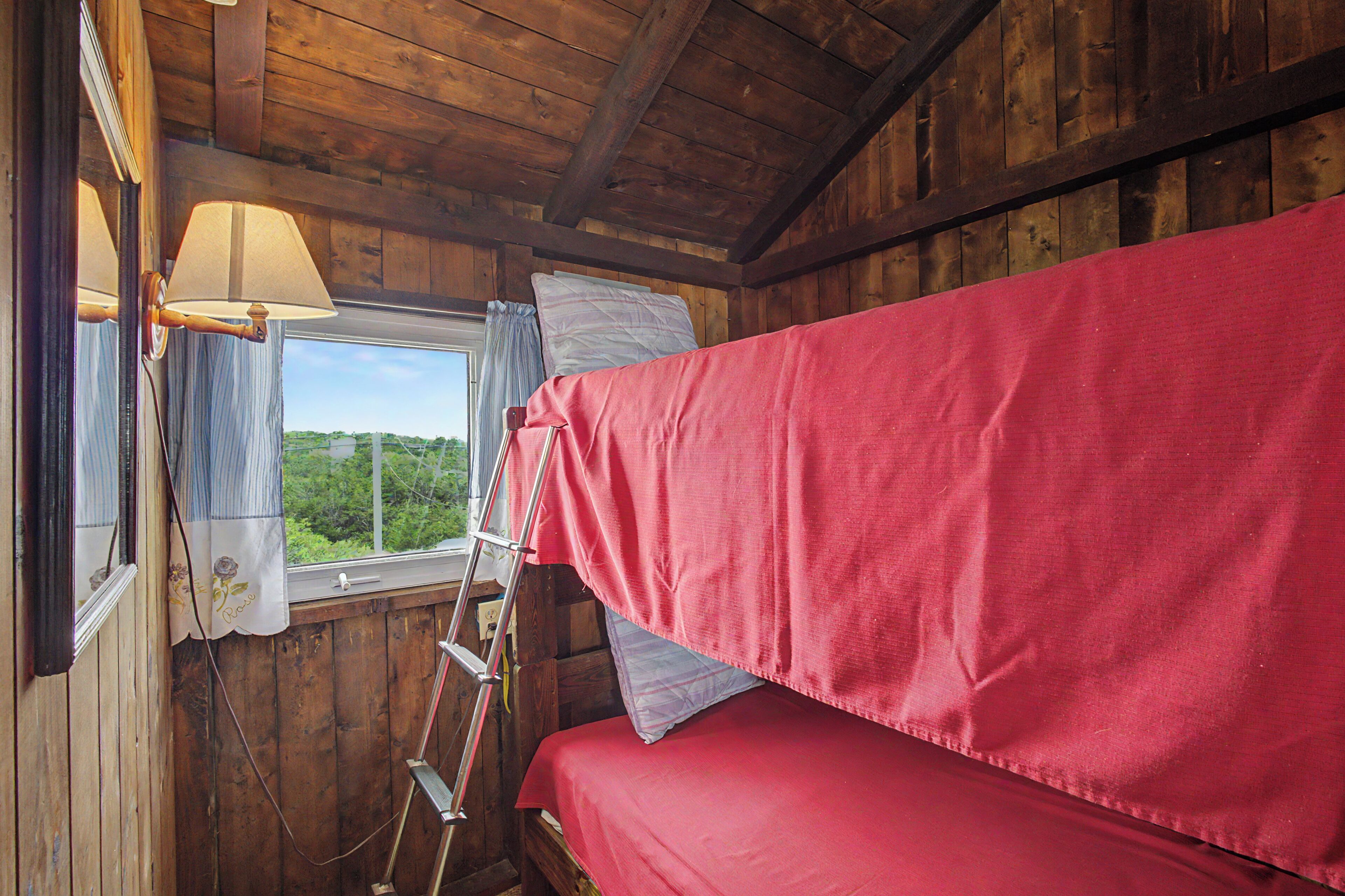 A room with wooden walls, a bunk bed with red bedding, a window with curtains, and a ladder leaning against the wall.