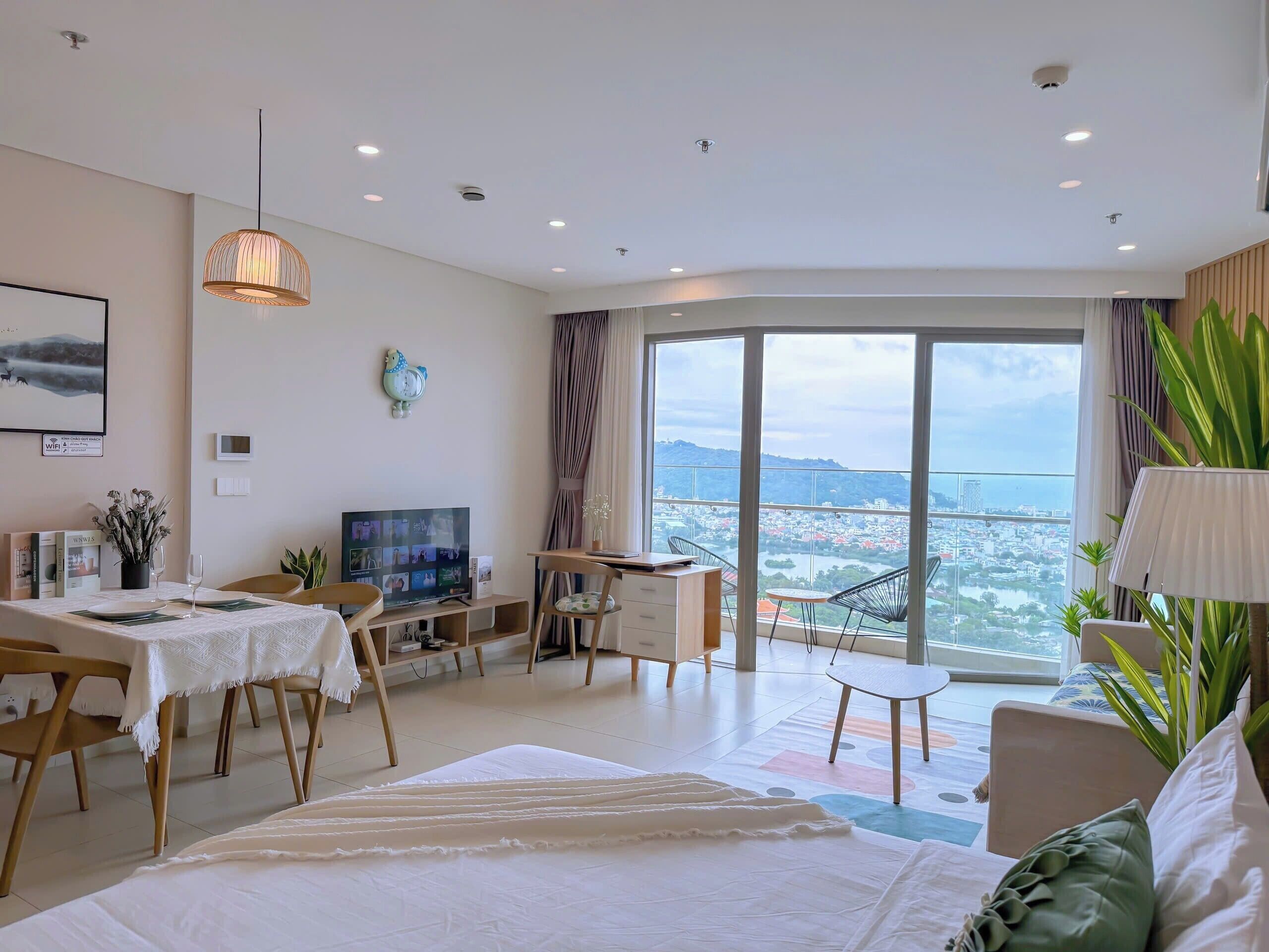 A modern hotel room with a large bed, a dining area, a TV, and a balcony with a city view.
