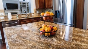 A kitchen with granite countertops, a stainless steel refrigerator, wooden cabinets, and a fruit basket on the counter. - Luxury Secluded Family Retreat (Apex)