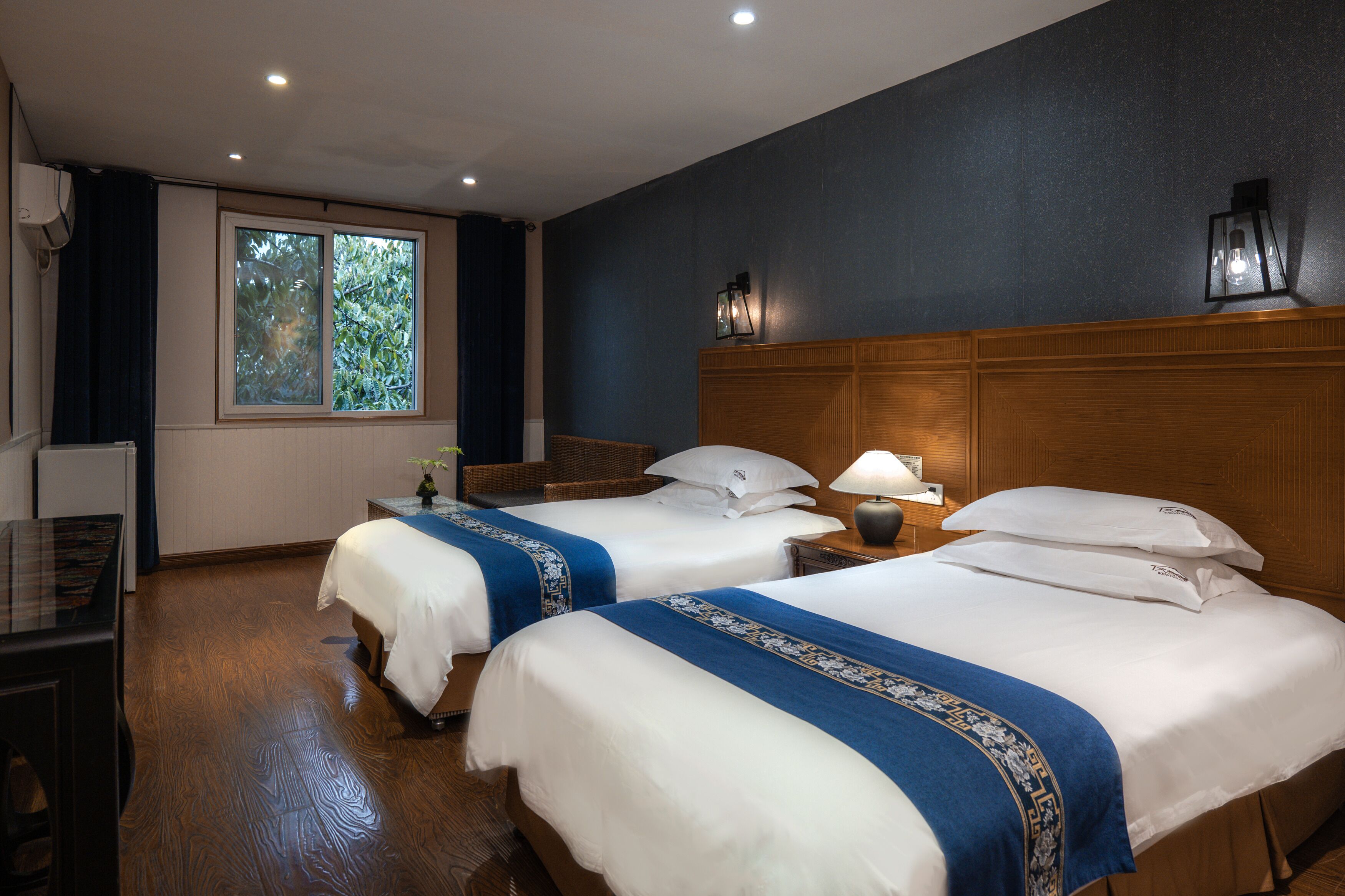 Habitación de hotel con dos camas, un escritorio, una ventana con cortinas y una mesita de noche con lámpara.