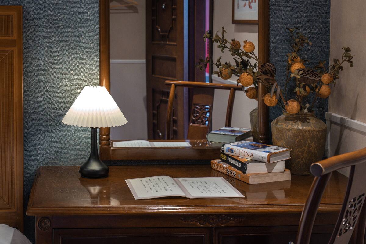 A wooden desk with a lamp, open book, and a vase with dried flowers.