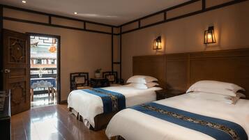 Habitación de hotel con dos camas, cabecera de madera, mesita de noche, una mesa con un florero y vista a un edificio tradicional a través de una puerta abierta.