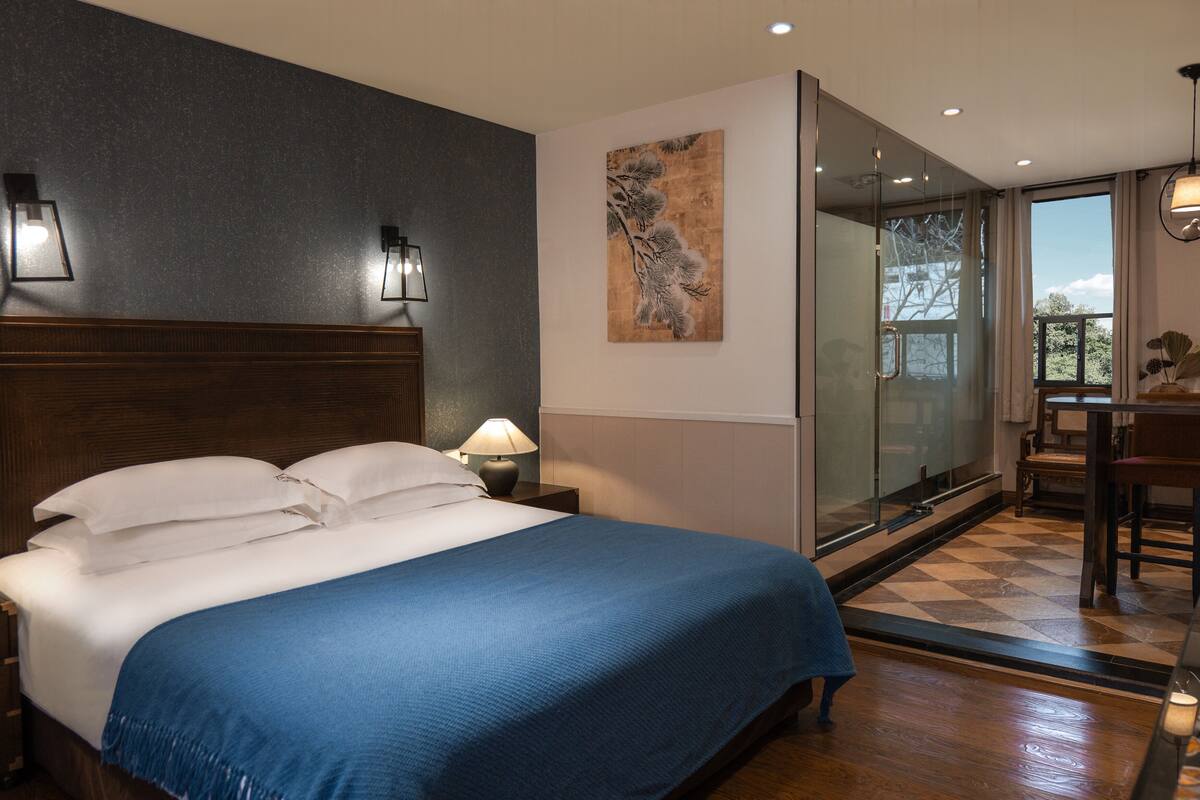 A bedroom with a large bed, a nightstand, a lamp, a painting, a glass door leading to a balcony, and a chair.