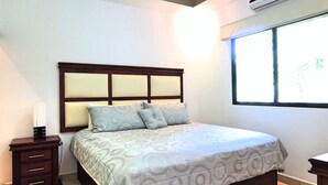 A neatly made bed with a patterned bedspread, a wooden headboard, and a bedside table with a lamp. - Hidden Palms Unit #12 (Nosara)